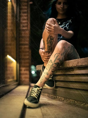 Woman displaying trendy calf tattoos with bold designs including a butterfly, sitting on steps in an urban setting. Temporary tattoos for legs.